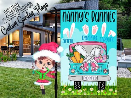 Nanny's Bunnies garden flag with named bunny-ear and paw motifs above a turquoise truck on blue outside a house.
