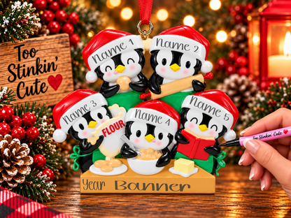 A festive Christmas ornament shaped like five penguins cooking together, with customizable name labels on their hats and a banner at the bottom. A hand is holding a pink marker to write on the ornament, set against a warm holiday background with lights, pinecones, and red ornaments.