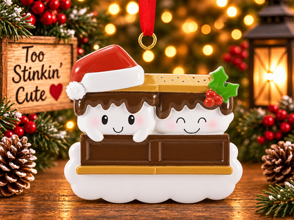 S’mores couple ornament hanging from a red ribbon on a Christmas tree beside a wooden Too Stinkin’ Cute sign.