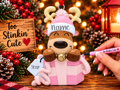 Smiling brown reindeer ornament with cream antlers, a pink pom-pom hat, and a gold-colored bell.