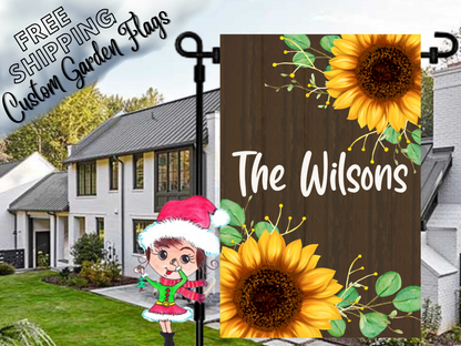 Garden flag mockup with bright yellow sunflowers and family-name lettering on a dark wood-look background.