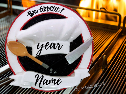 Christmas Ornament showing white chef hat with wooden spoon and knife and red-and-black circular background.